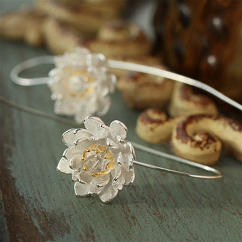 Boucles d’oreilles argentées en forme de fleur de lotus avec des détails dorés, posées sur un fond bleu patiné.