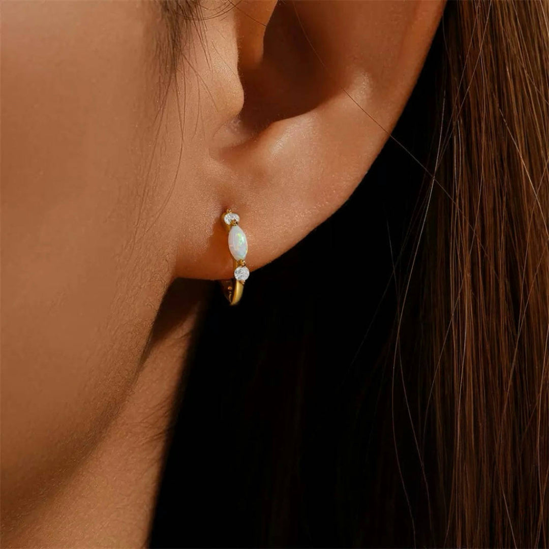 Boucles d'oreilles créoles dorées ornées d'une opale ovale blanche et de deux petits diamants brillants, portées à l'oreille.