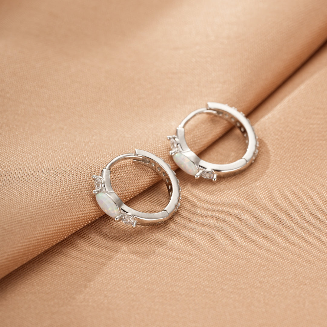 Paire de boucles d'oreilles créoles en argent ornées d'opales ovales et de petits cristaux scintillants sur fond satiné beige.
