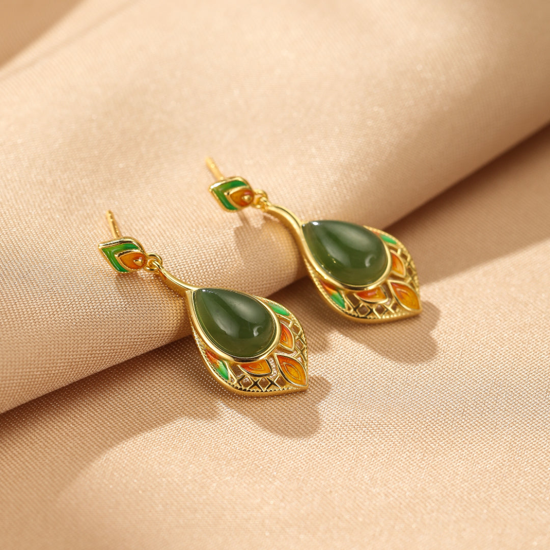 Paire de boucles d'oreilles pendantes en jade vert émeraude, plaqué or, avec des détails émaillés colorés formant des feuilles et des motifs floraux sur un fond beige satiné.
