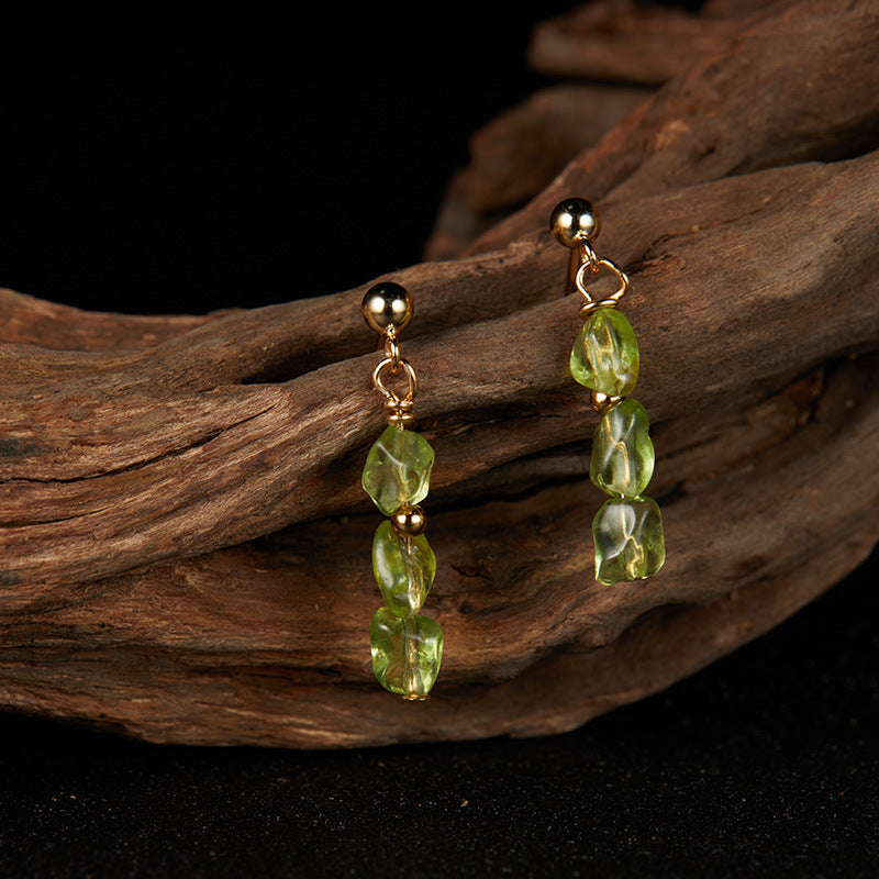 Boucles d’oreilles pendantes en argent plaqué or avec des pierres de péridot naturelles brutes en forme de goutte, sur fond sombre et bois.