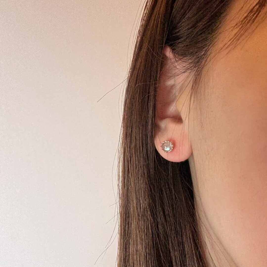 Boucle d'oreille en argent 925 plaqué or avec une pierre ronde facettée type péridot ou labradorite, mise en valeur par un entourage texturé, portée à l'oreille d'une femme aux cheveux bruns.