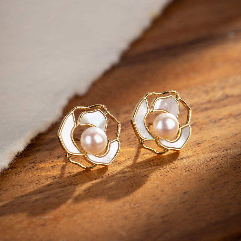 Boucles d'oreilles plaqué or en forme de rose avec perle nacrée au centre sur fond de bois.