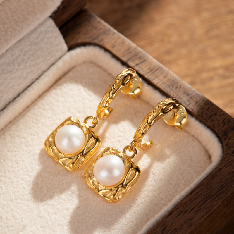 Boucles d'oreilles pendantes en argent plaqué or avec une perle blanche naturelle enchâssée dans un cadre carré texturé.