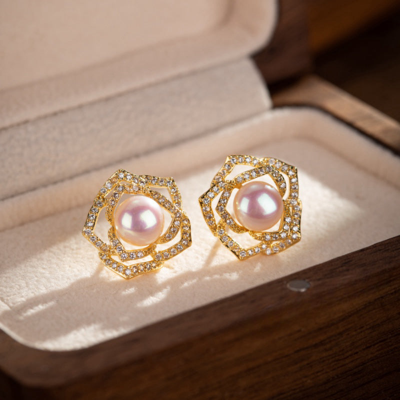 Boucles d'oreilles en forme de rose en or plaqué serties d'une perle rose et de petits diamants scintillants, présentées dans une boîte à bijoux.