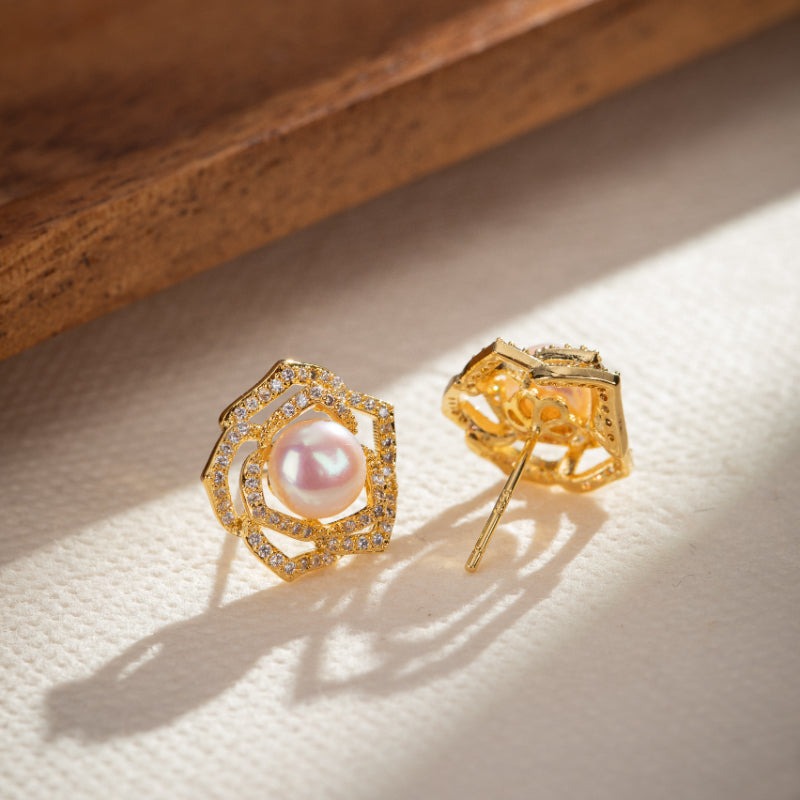 Boucles d'oreilles en forme de rose ajourée en argent plaqué or, serties de petites pierres scintillantes et ornées d'une perle rosée au centre.