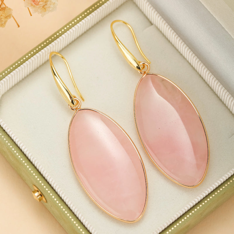 Paire de boucles d'oreilles pendantes ovales en quartz rose montées sur une monture dorée, présentées dans un écrin vert.
