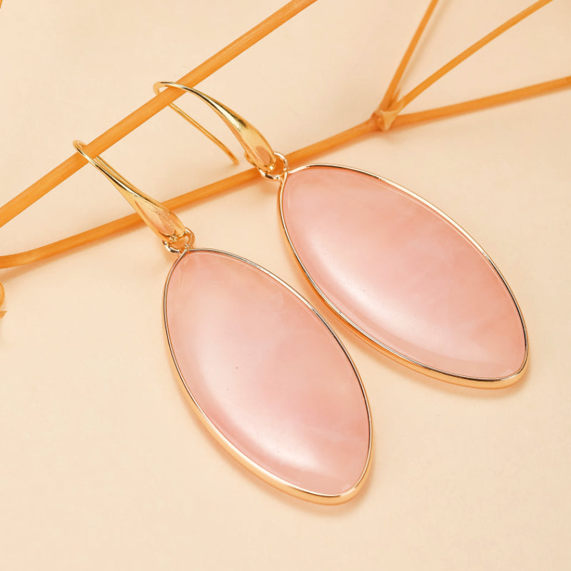 Boucles d'oreilles pendantes avec une pierre de quartz rose ovale et une monture dorée sur fond beige.