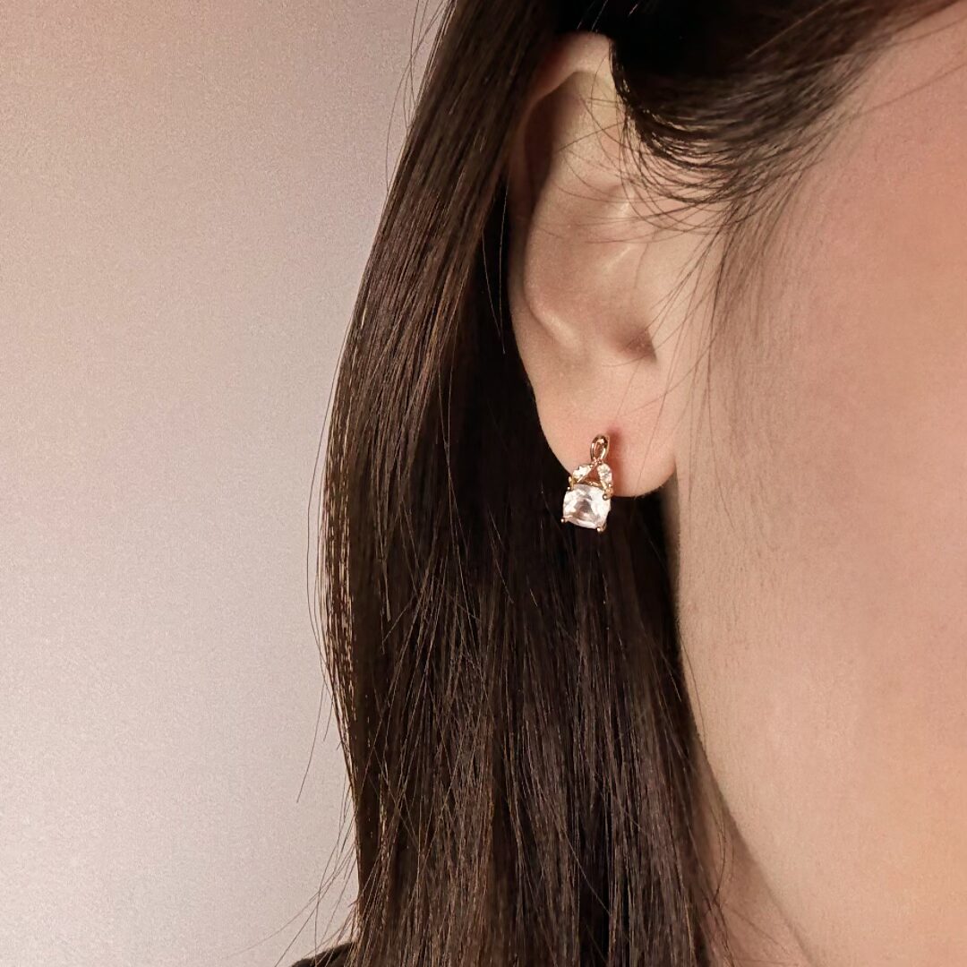 Boucles d'oreilles pendantes en argent plaqué or ornées d'une pierre carrée transparente et de petites pierres lumineuses, portées à l'oreille d'une femme.