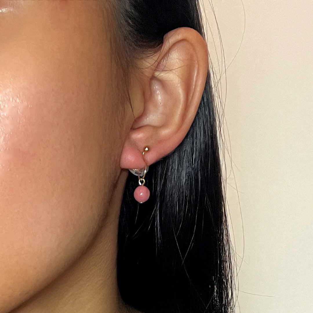 Boucles d’oreilles créoles argentées avec une perle rose de rhodonite suspendue, portées par une femme.