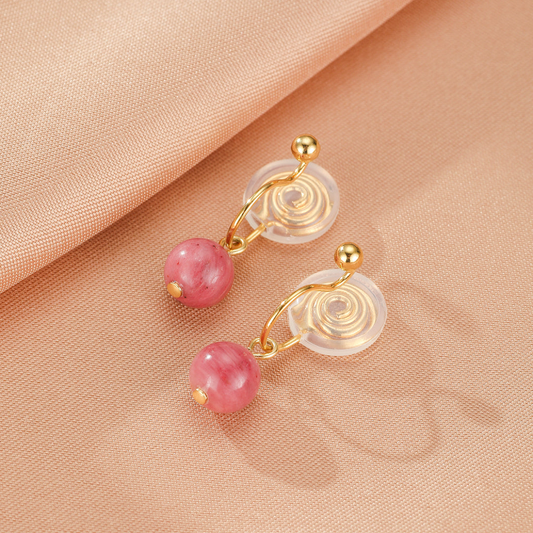 Paire de boucles d'oreilles pendantes avec des perles rondes en rhodonite rose sur un fond de tissu satiné pêche.