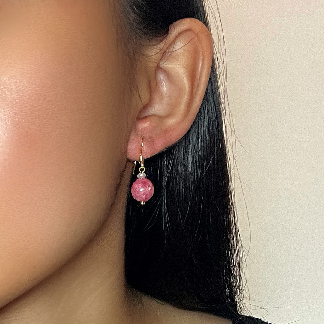 Une femme porte des boucles d'oreilles pendantes en or plaqué, ornées d'une perle sphérique rose vif et d'une petite perle blanche.