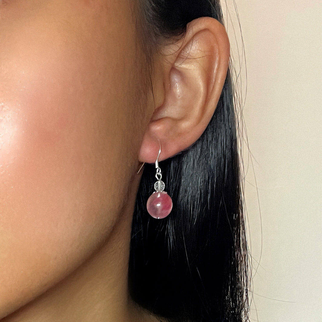 Gros plan d'une boucle d'oreille en argent avec une grosse perle ronde rose de rhodonite et une petite perle transparente au-dessus, portée à l'oreille d'une femme.