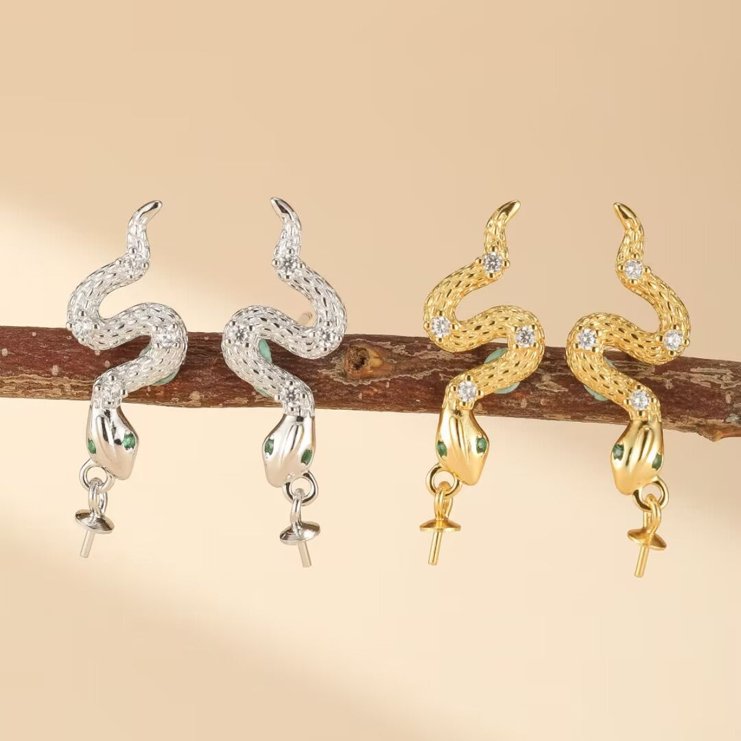Deux paires de boucles d'oreilles en forme de serpent, l'une en argent 925 sertie de zircons et d'émeraudes, l'autre dorée ornée de zircons et d'émeraudes, présentées sur une branche.