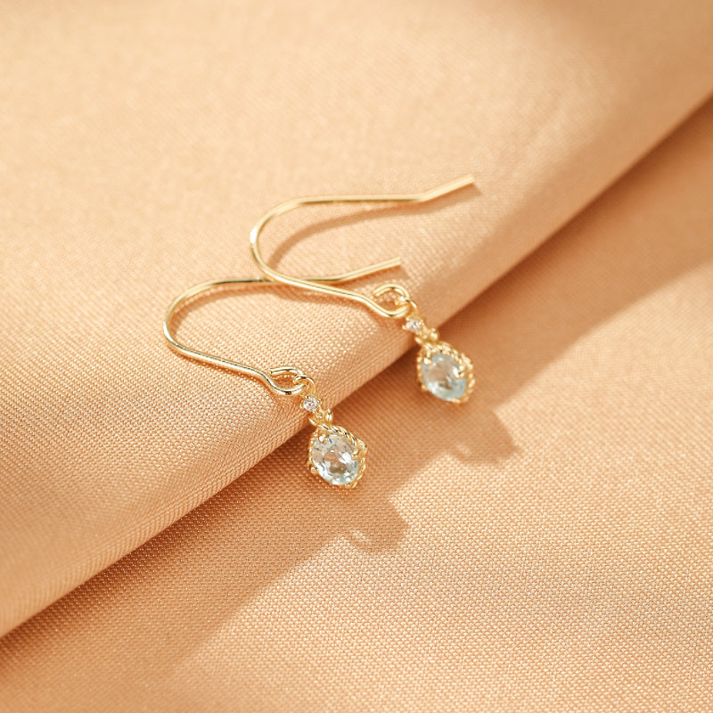 Boucles d'oreilles pendantes en argent plaqué or avec topazes bleues ovales et petits diamants scintillants, sur fond texturé couleur pêche.