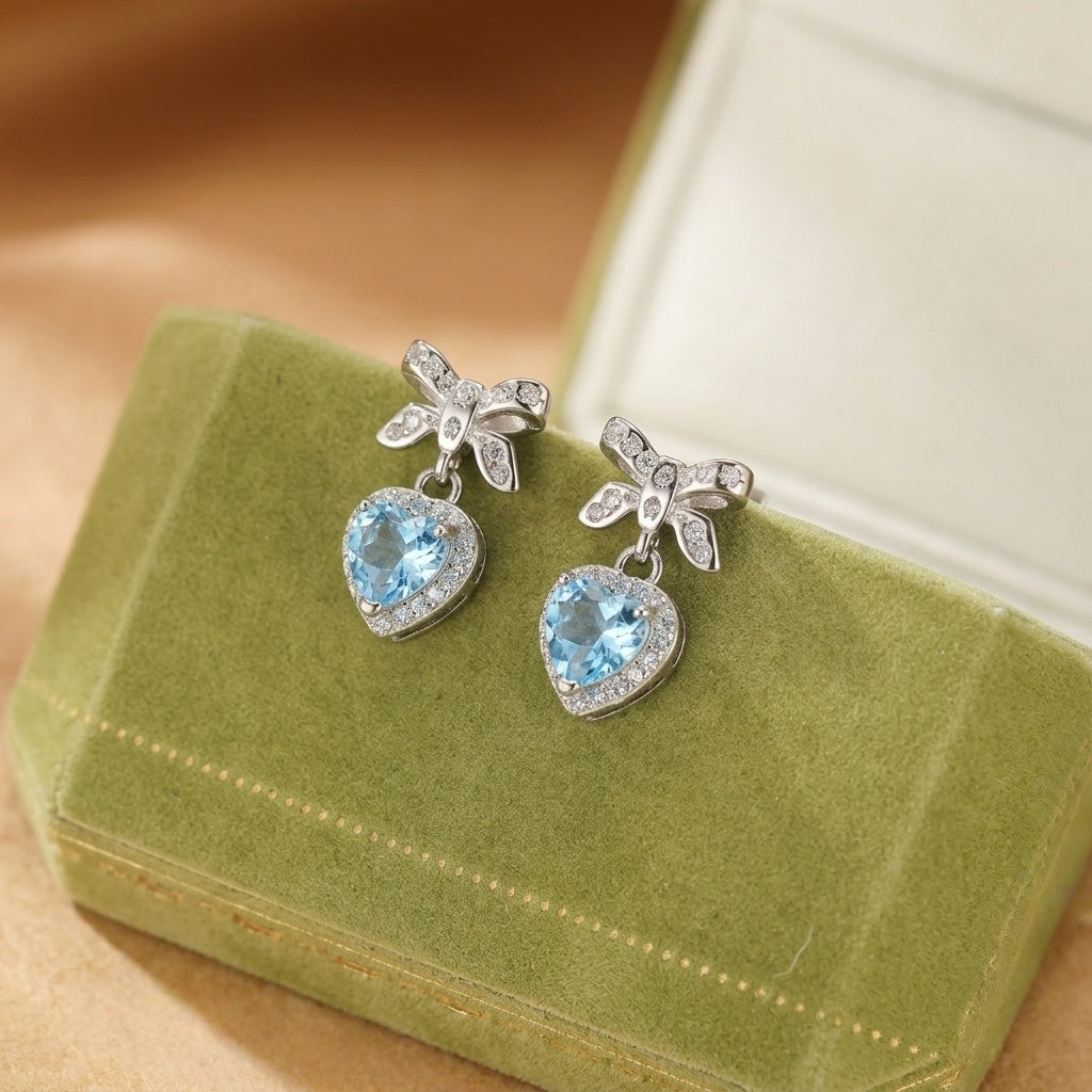 Boucles d'oreilles cœur en topaze bleue serties de diamants avec des nœuds en argent, présentées sur une boîte à bijoux verte.