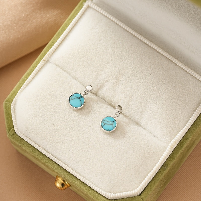 Boucles d'oreilles Aura du Lagon en argent et plaqué or avec une turquoise bleue ronde et veinée, présentées dans un écrin vert.