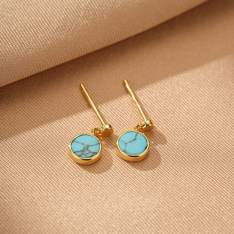 Paire de boucles d'oreilles pendantes en argent plaqué or avec une turquoise ronde aux veines bleues et grises, sur fond beige texturé.