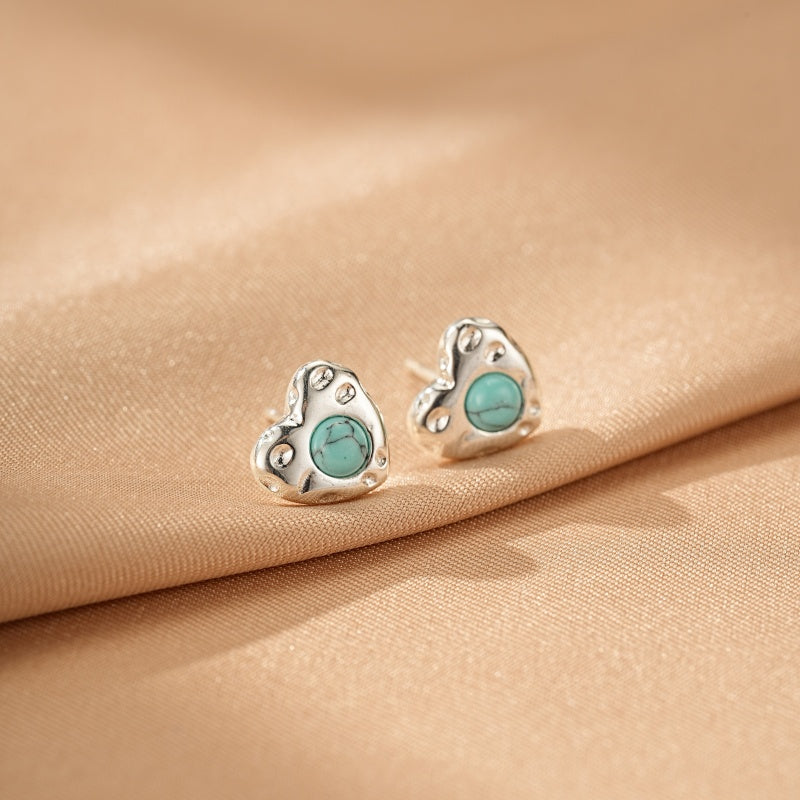 Boucles d'oreilles en argent en forme de cœur avec une turquoise au centre, posées sur un tissu satiné beige.