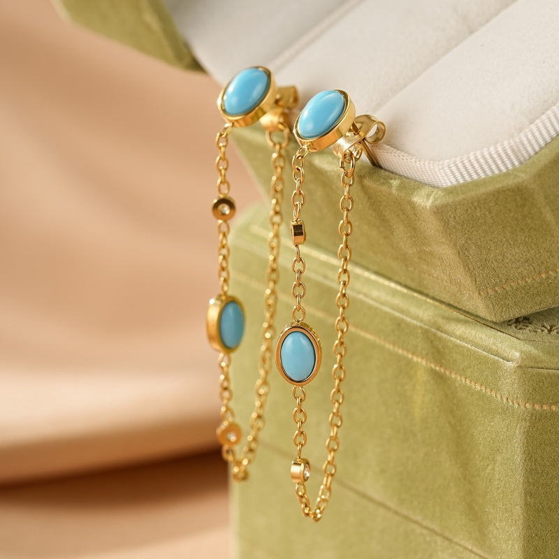 Boucles d'oreilles pendantes en or avec des turquoises ovales et des petits cristaux, présentées dans une boîte à bijoux verte.