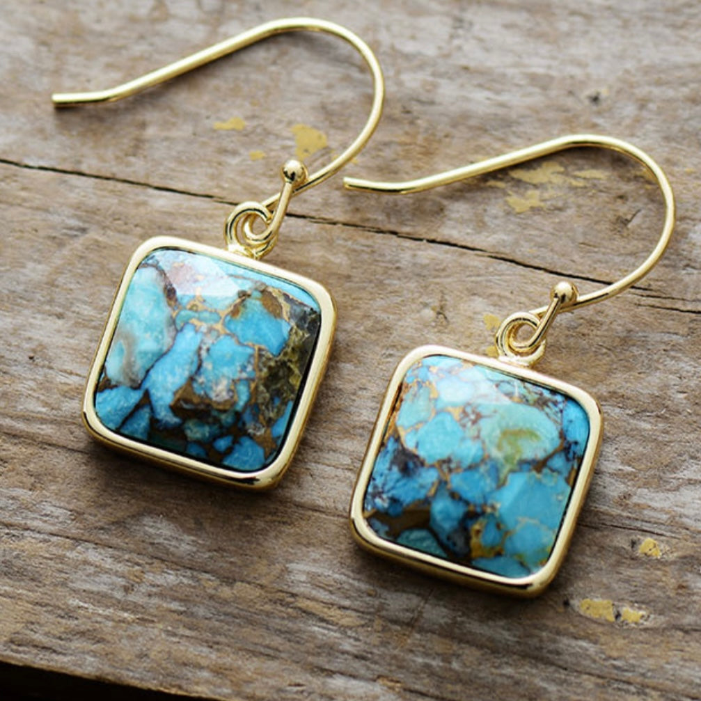 Boucles d'oreilles pendantes en argent plaqué or avec une pierre carrée turquoise veinée de brun, sur fond de vieux planches de bois