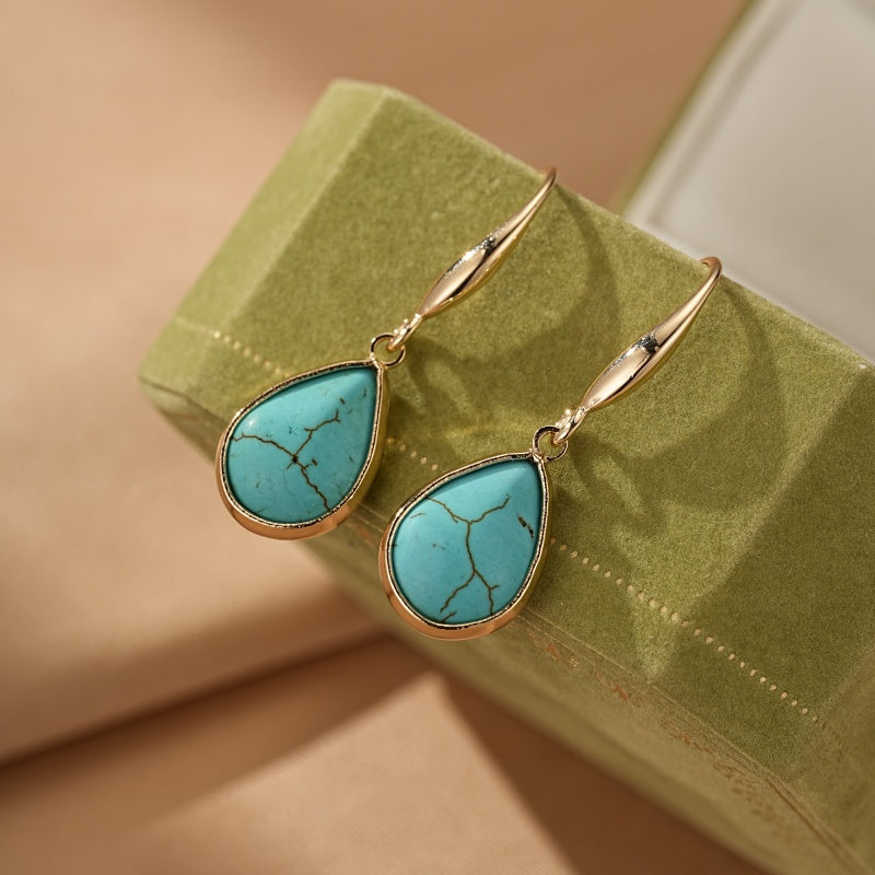 Boucles d'oreilles pendantes dorées avec des pierres turquoise en forme de goutte, sur un fond vert et beige.