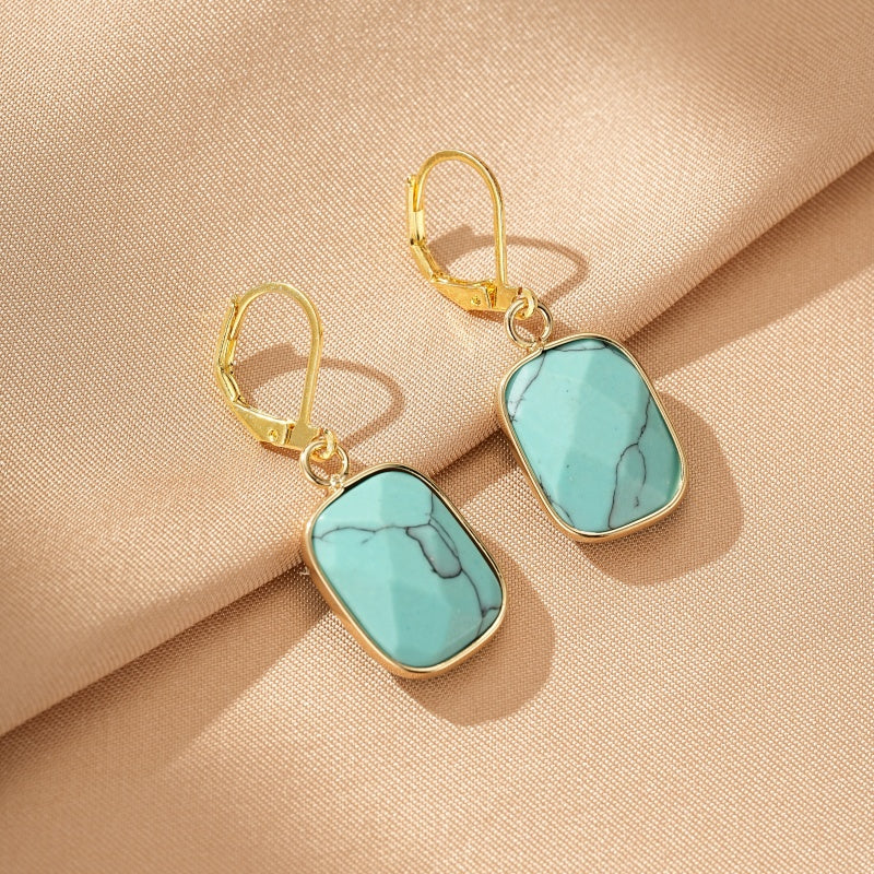Boucles d'oreilles rectangulaires plaquées or avec pierre turquoise facettée et veinures noires, sur fond beige.