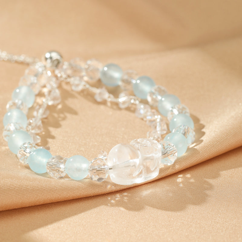 Bracelet double rang fait de perles d'aigue-marine bleutées et de cristaux de roche transparents facettés, avec une perle centrale sculptée en forme de crâne en cristal de roche, sur un fond en tissu satiné beige.