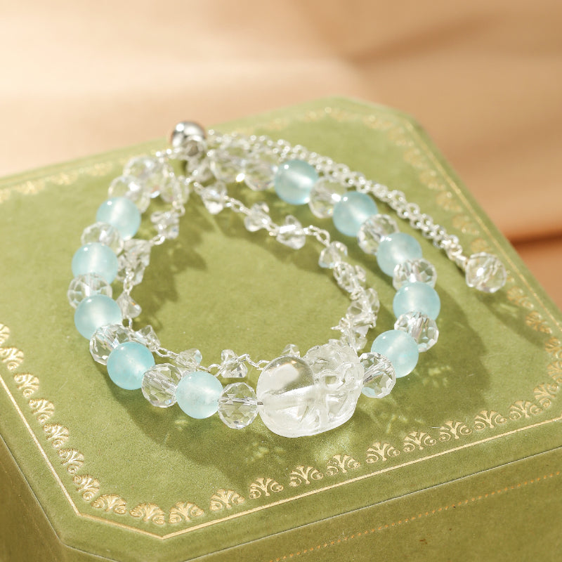 Bracelet multicouche en aigue-marine et cristal de roche, avec un fermoir en alliage argenté. Ce bijou délicat est posé sur une boîte verte ornée.