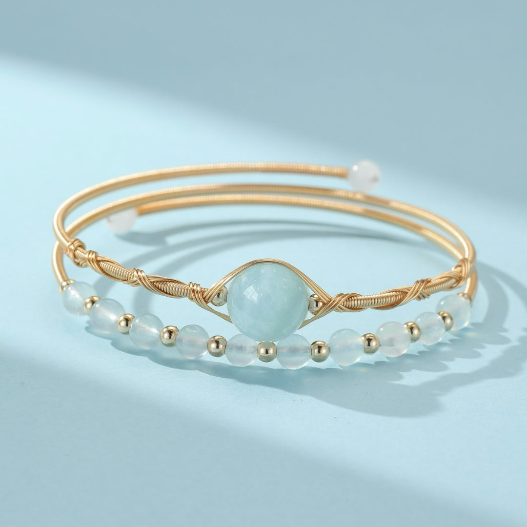 Ensemble de deux bracelets fins dorés ornés de perles bleu clair et d'une grande perle centrale d'aigue-marine, sur fond bleu pastel.
