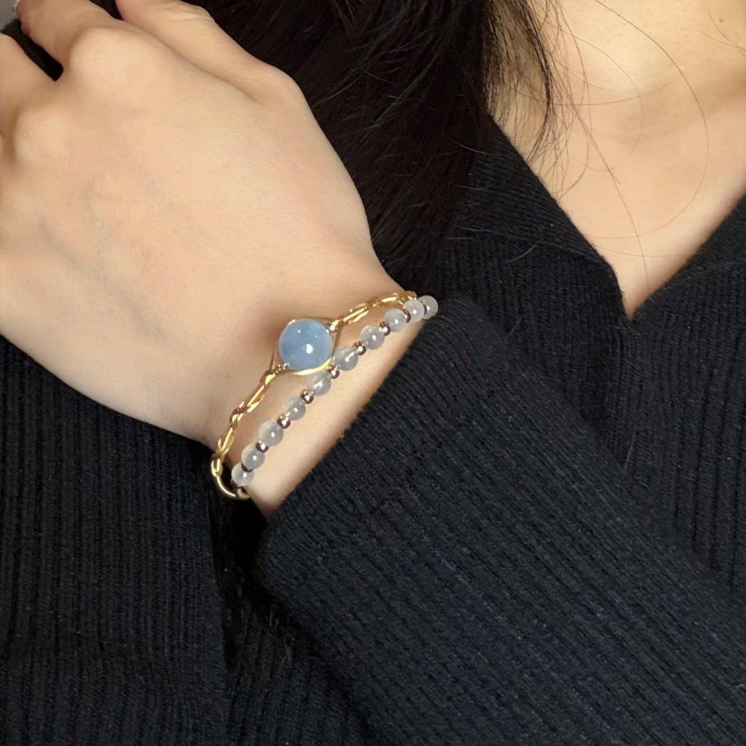 Gros plan sur un bracelet fin en or avec une perle bleue ciel centrale et un bracelet de petites perles grises porté au poignet d'une femme