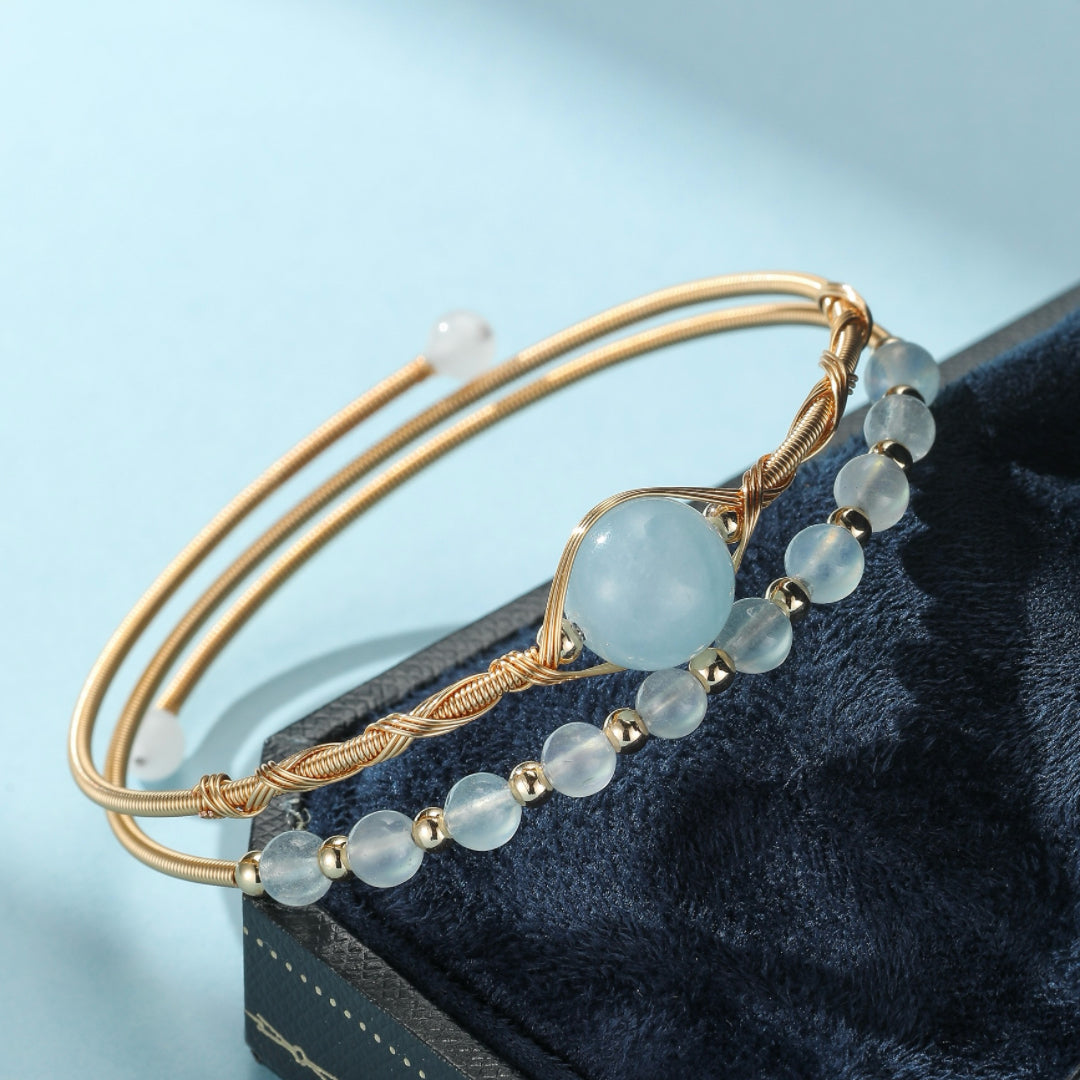 Bracelet jonc doré avec une grosse perle bleue d'aigue-marine au centre, entourée de petites perles bleu clair et de perles dorées. Le bracelet est ouvert et repose dans une boîte bleue.