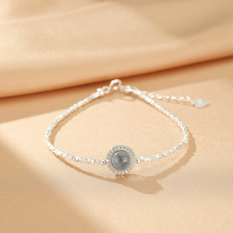 Bracelet fin en argent avec une pierre d'aigue-marine ronde entourée de petits cristaux, sur fond beige satiné.