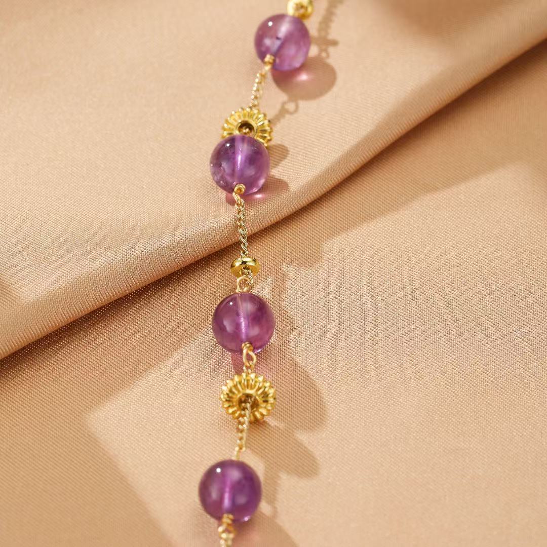 Bracelet en or fin avec des perles d'améthyste violettes et des ornements dorés en forme de fleur, sur un fond de tissu satiné beige.