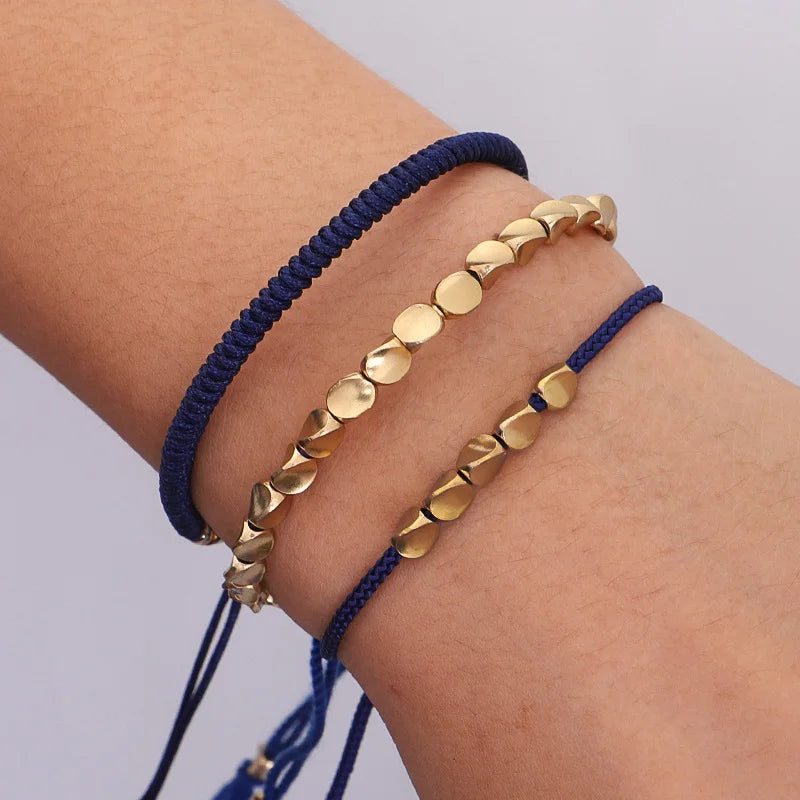 Trois bracelets fins en cordon bleu marine avec des perles dorées en forme de disque et de cube, vue rapprochée sur un poignet.