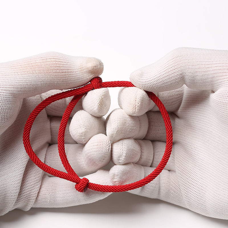 Gros plan d'un bracelet minimaliste en cordon rouge noué, tenu par des mains gantées de blanc. Symbole de force, idéal pour couple ou amis.