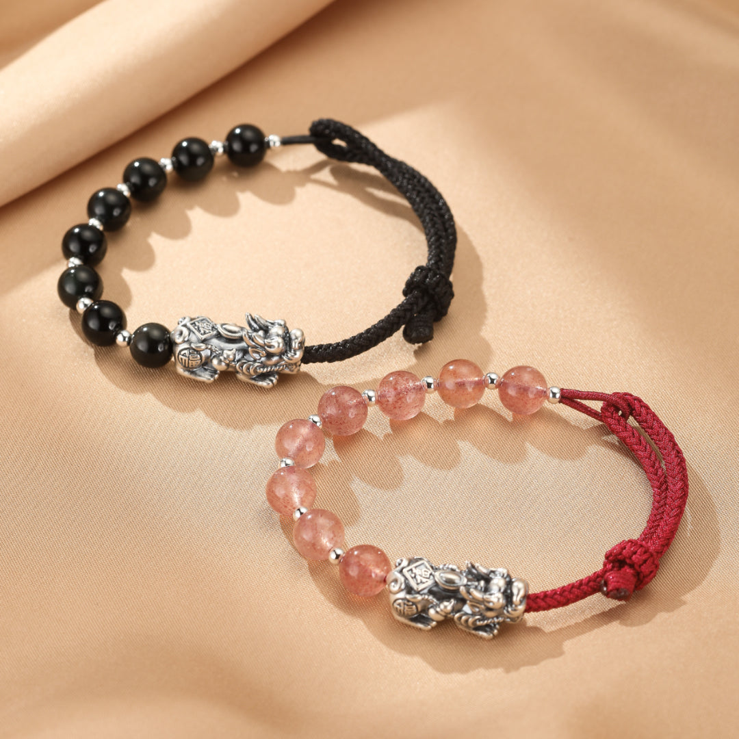 Deux bracelets assortis pour couple, l'un avec des perles noires et un Pi Xiu en argent, l'autre avec des perles roses et un Pi Xiu en argent, reliés par un fil rouge.