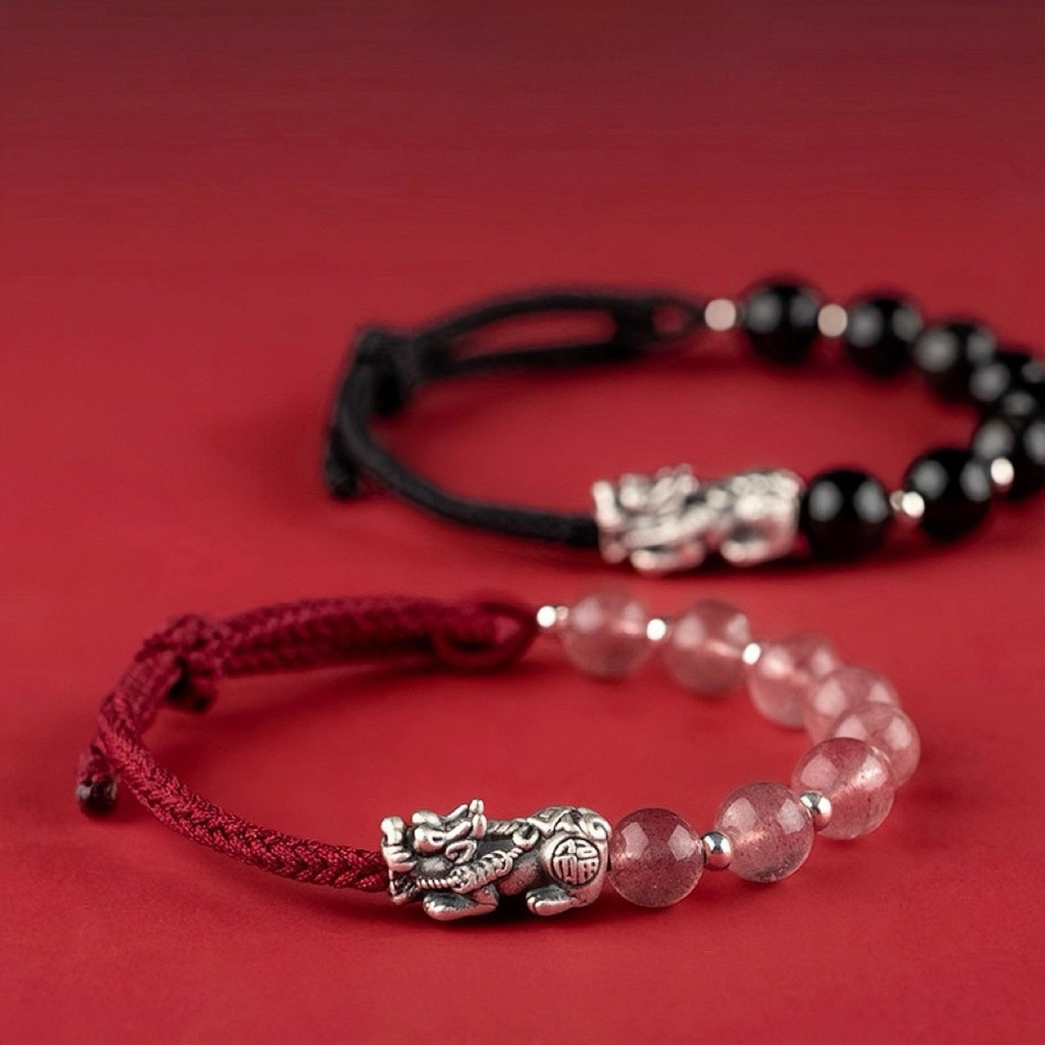 Deux bracelets assortis pour couple, l'un avec des perles noires et l'autre avec des perles roses, ornés chacun d'une breloque Pi Xiu en argent sur un fil rouge ou noir.