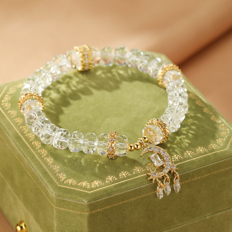 Bracelet en cristal de roche avec un pendentif en forme de croissant de lune et d'étoiles, orné de zircons et de perles dorées, posé sur une boîte verte.