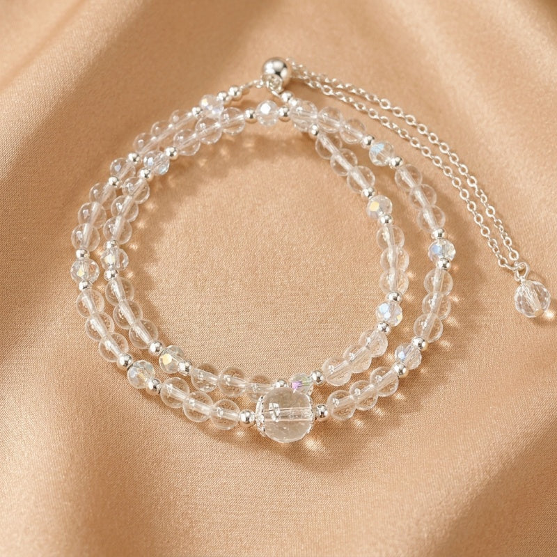 Bracelet double rang en cristal de roche transparent et perles argentées avec un fermoir réglable, sur un fond satiné beige.