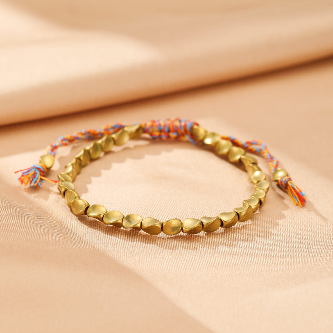 Bracelet en cuivre avec des perles dorées texturées en forme de cœur, reliées par un cordon tressé multicolore aux extrémités avec des pompons.