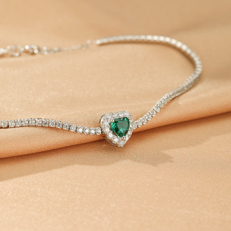 Bracelet fin en argent 925 orné d'un pendentif en forme de cœur vert émeraude entouré de petits diamants scintillants, posé sur un tissu satiné beige.