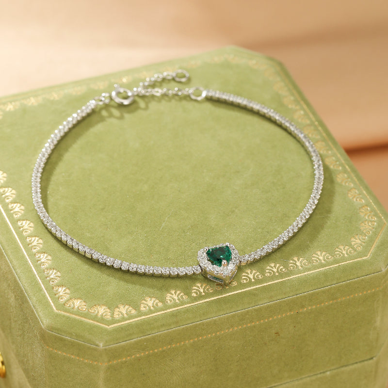 Bracelet fin en argent 925 avec un pendentif émeraude en forme de cœur, entouré de petites pierres scintillantes, posé sur une boîte verte.