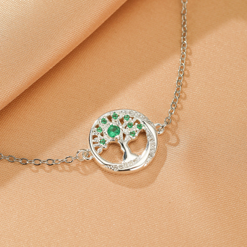 Bracelet en argent avec un pendentif arbre de vie rond orné d'émeraudes et de zircons cubiques, le tout sur un fond de tissu brun clair.