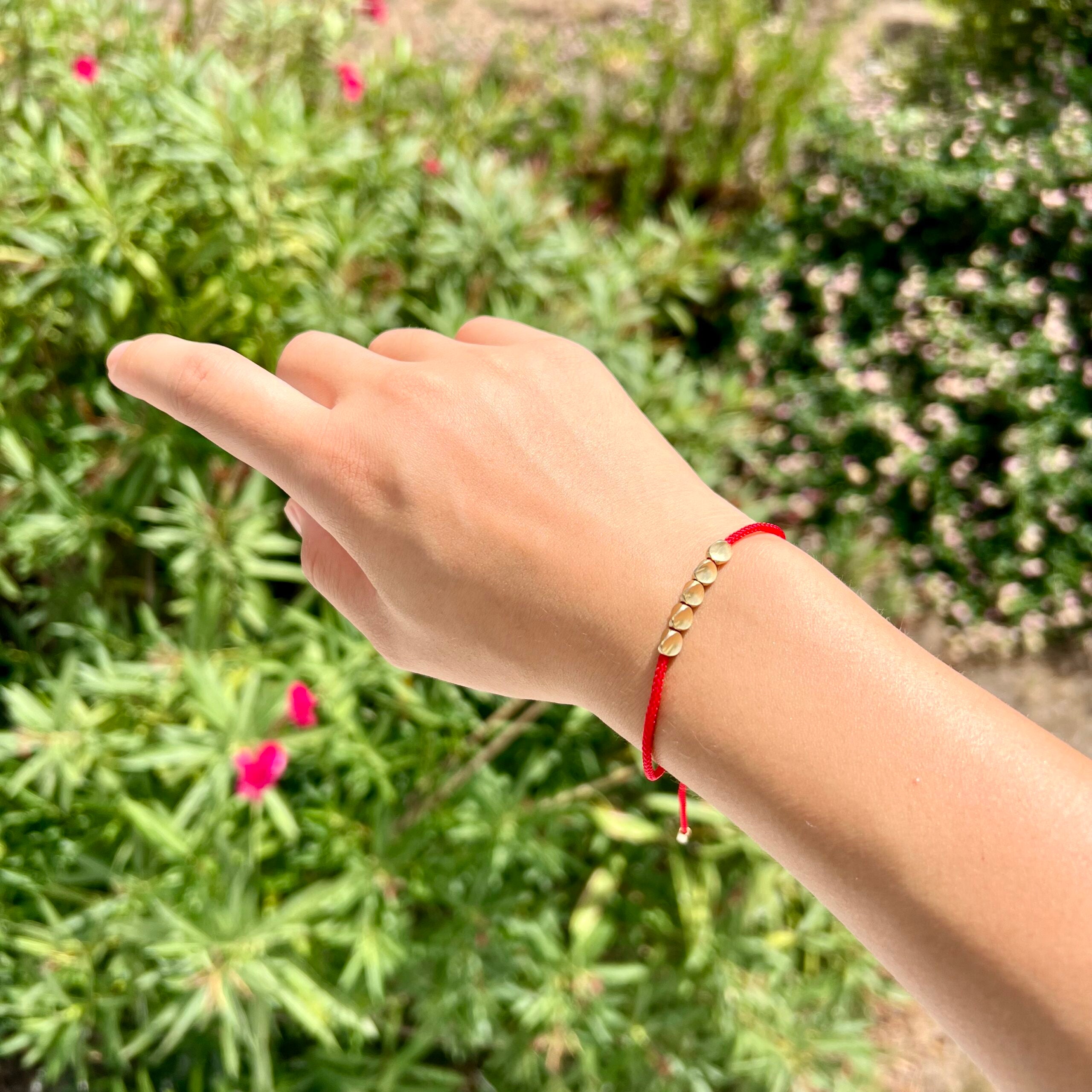 Gros plan d'un bracelet fil rouge avec des perles dorées sur le poignet d'une main, avec un arrière-plan flou de verdure.