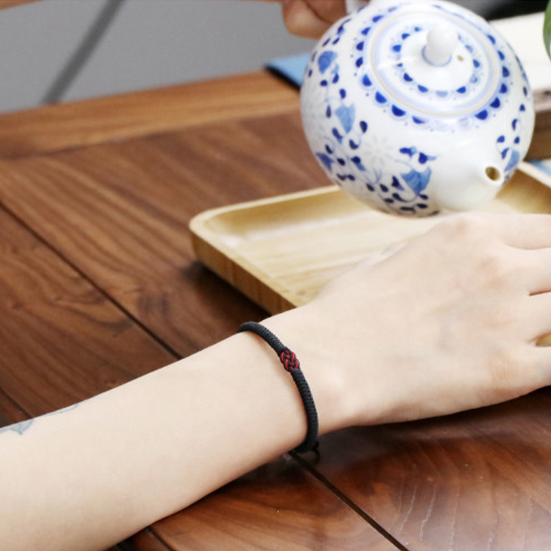 Bracelet fil rouge tressé noir avec un nœud central rouge, porté au poignet gauche, posé sur une table en bois, à côté d'une théière en céramique bleue et blanche.