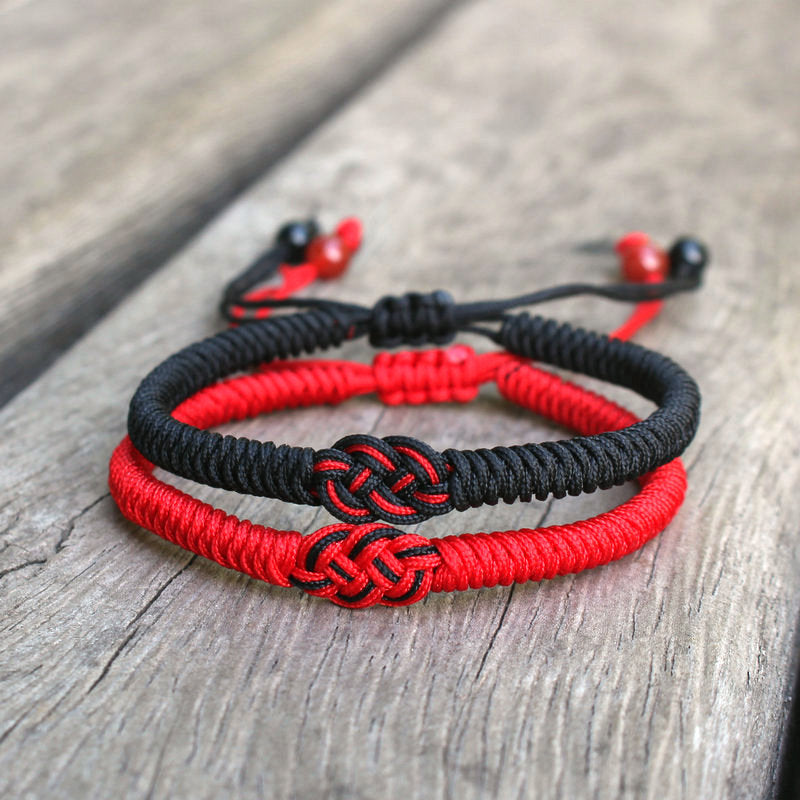 Deux bracelets tressés, un rouge et un noir, avec un nœud central complexe, sur un fond en bois.