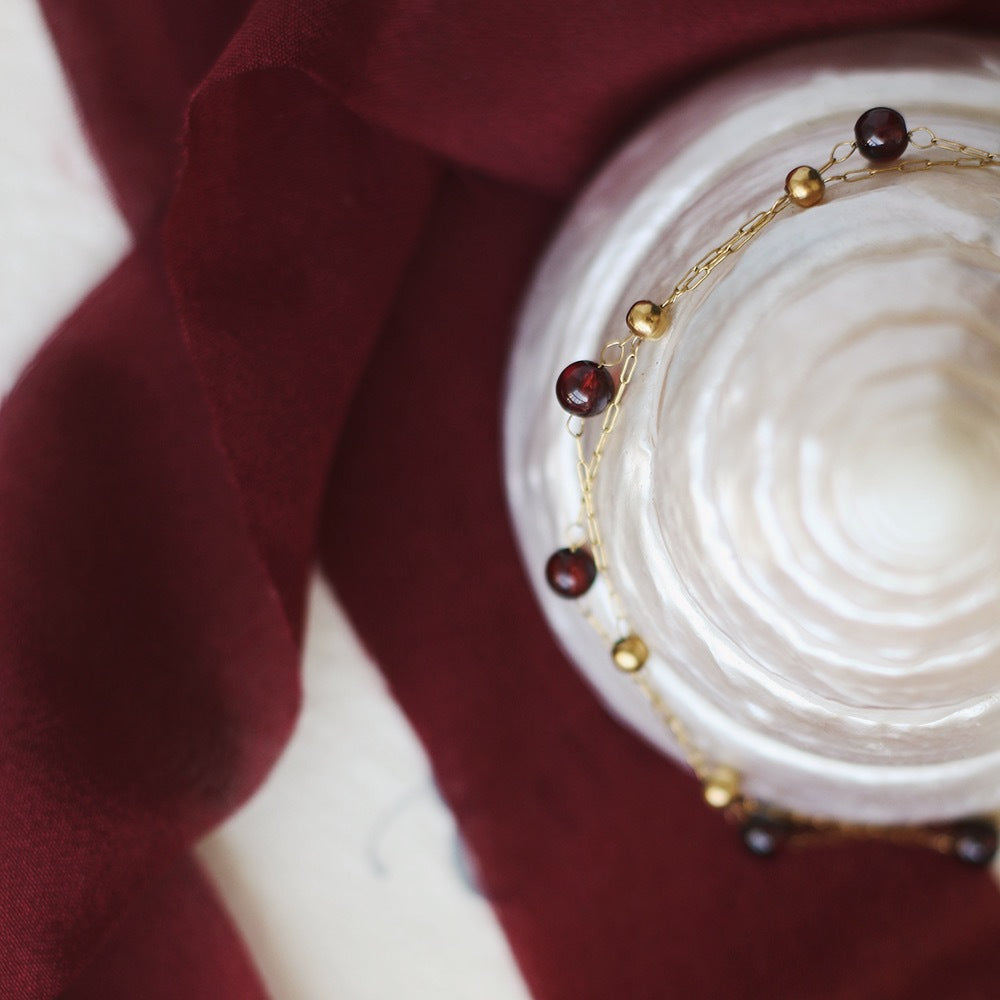 Bracelet fin en plaqué or 18K, orné de perles de grenat rouge foncé et de petites perles dorées, posé sur une surface nacrée avec un drapé bordeaux