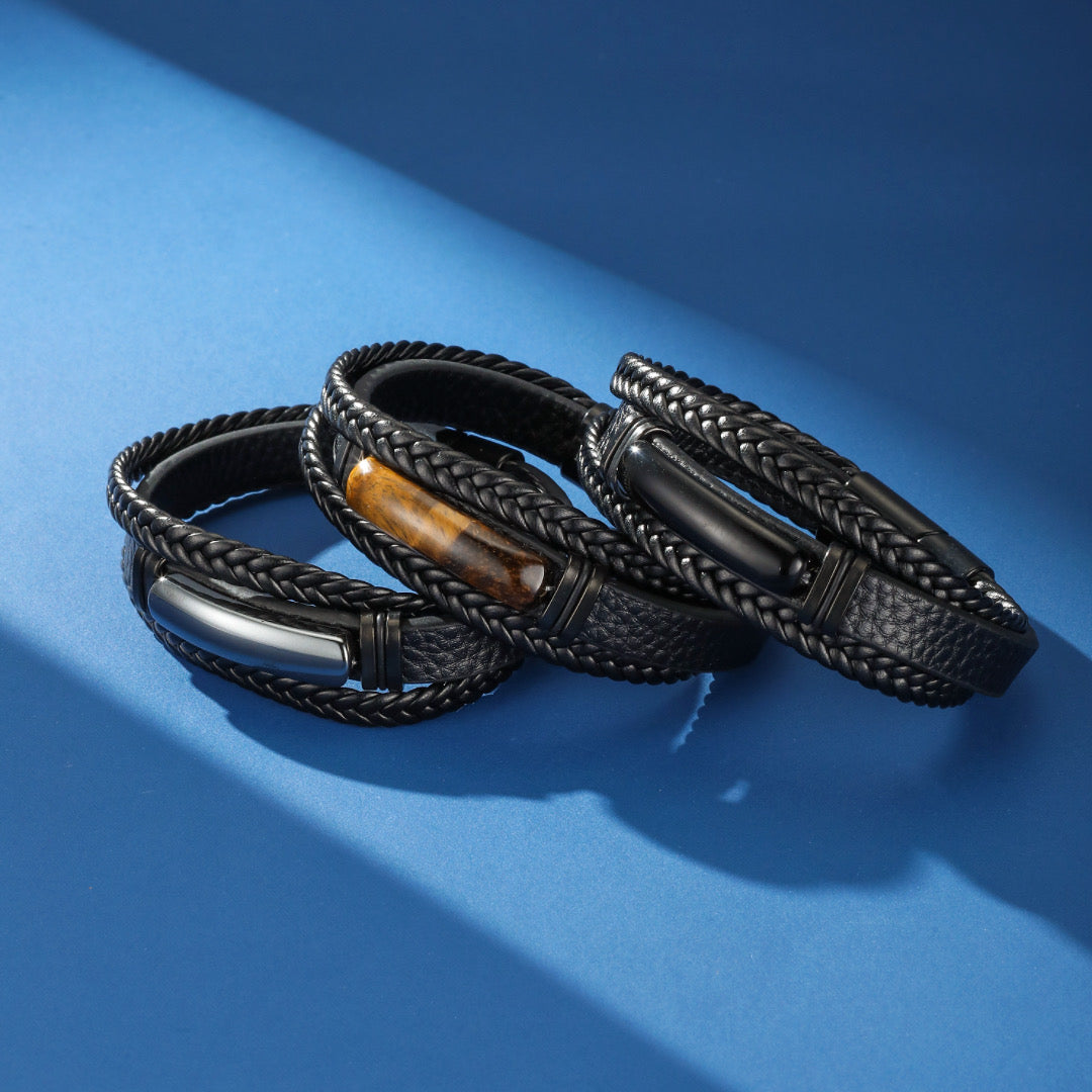 Trois bracelets homme en cuir tressé noir, ornés d'une barre métallique argentée, d'une pierre œil de tigre et d'une barre noire mate, sur fond bleu.