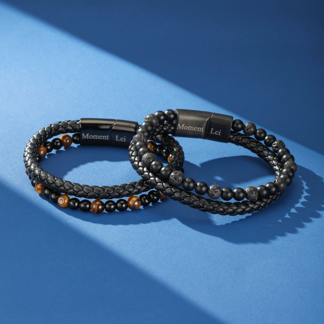 Deux bracelets pour homme en cuir tressé noir, ornés de perles d'œil de tigre et de pierre de lave, avec un fermoir magnétique gravé du logo 'Moment Lci'.
